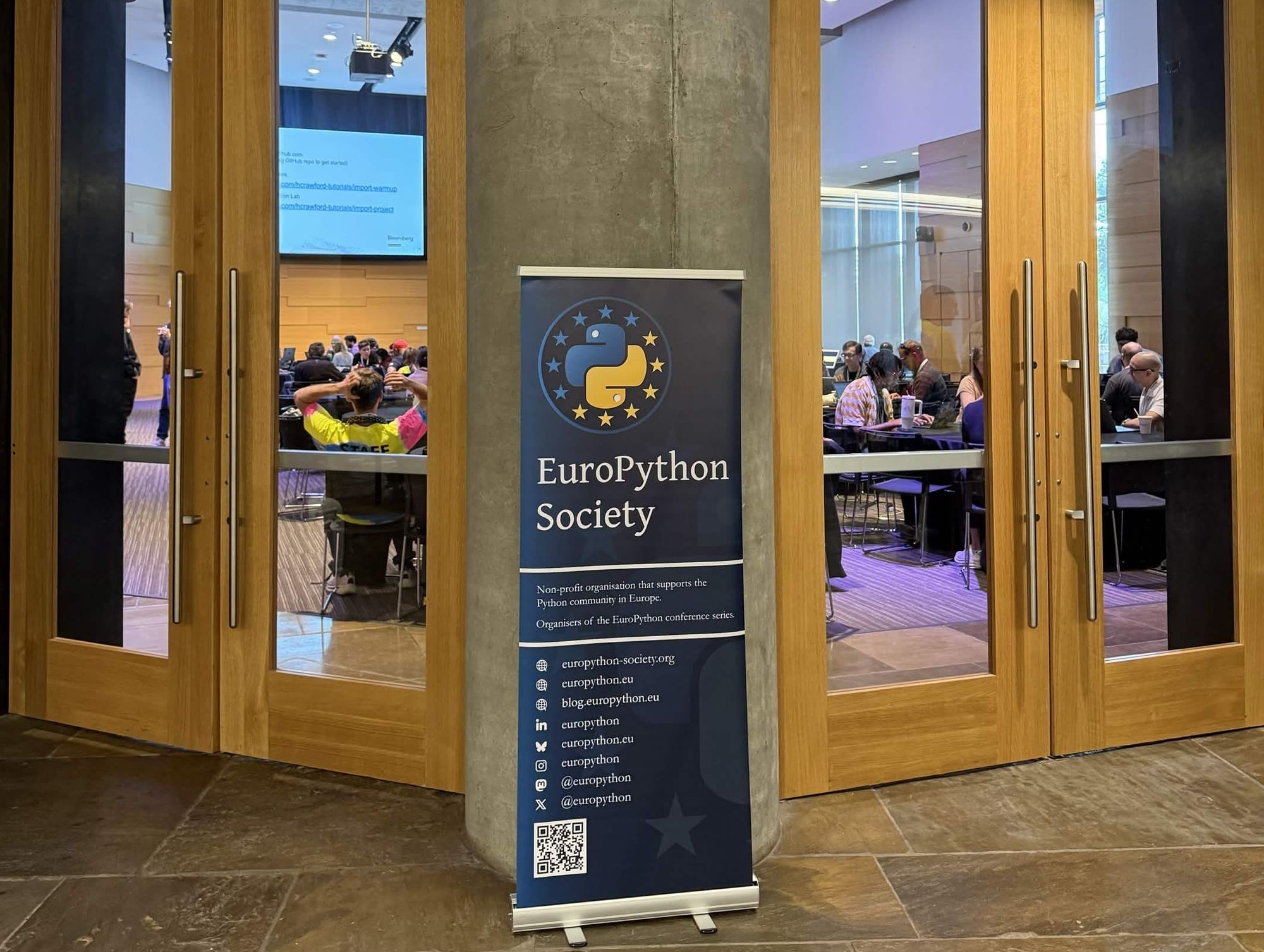 A photo of a EuroPython Society banner in an event venue, with wooden glass doors through which attendees can be seen